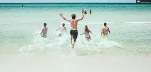 A group of people running into the ocean on a sunny beach day, with clear blue water and sky.