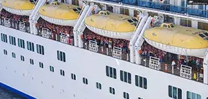 Cruise ship passengers in life vests during a lifeboat drill, with yellow lifeboats above them.