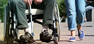 A person in a wheelchair holding a cane, accompanied by someone walking beside them outdoors