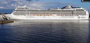 A large MSC cruise ship docked at a port, with a cloudy sky and calm water in the background.