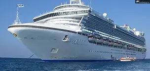 Large cruise ship docked near a tropical island with turquoise waters, lush greenery, and palm trees