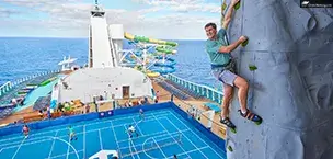 A smiling man climbs Star of the seas Rock Wall with the backdrop of a basketball court.