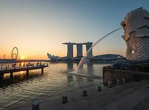 The iconic Merlion statue stands tall against a vibrant sunrise sky, with the city skyline reflected in the water below.