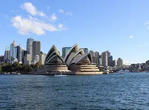 An iconic view of the Sydney Opera House, showcasing its distinctive sail-shaped roofs against a clear blue sky with the Sydney Harbour Bridge in the background.