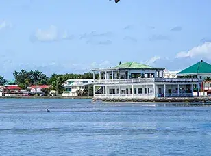 A bustling waterfront scene in Belize City. The image shows colorful buildings lining a waterway, with boats docked alongside. A large cruise ship is visible in the background.