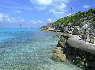 A white lighthouse stands on a rocky promontory overlooking a turquoise Caribbean sea. Lush greenery surrounds the lighthouse, and a sandy beach stretches out in the distance.