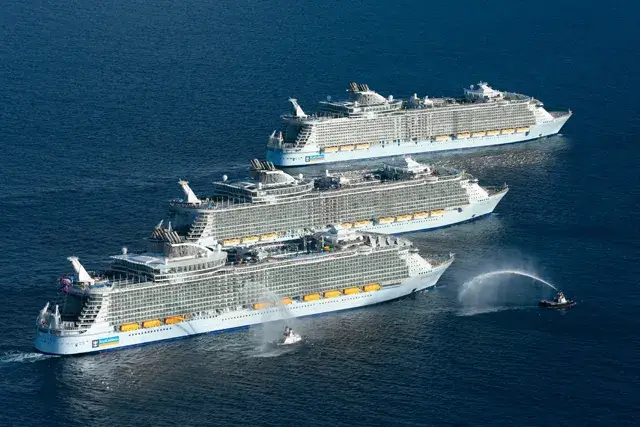 Harmony of the Seas, Allure of the Seas, and Oasis of the Seas are three large cruise ships side-by-side in the ocean. The ships are white with blue and yellow accents.