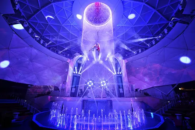 Icon of the Seas is a wide shot of two aerial artists and performers high above the stage in an AquaTheater show. Aqua Action, bubbling water fountains, excitement, daring, purple colors, stage lights