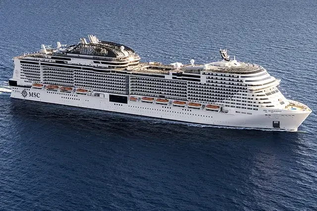 A large white cruise ship, the MSC Meraviglia, sails through calm blue waters with mountains in the distance.