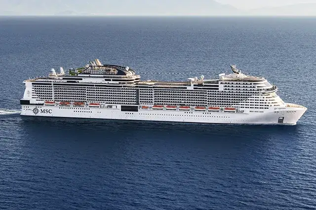 The MSC Virtuosa cruise ship sailing across calm blue waters with distant mountains in the background under a clear sky.