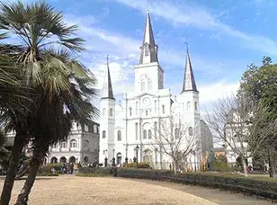 St. Louis Cathedral in Jackson Square, New Orleans. The iconic white cathedral with three spires stands in the center of the square, surrounded by palm trees and historic buildings.