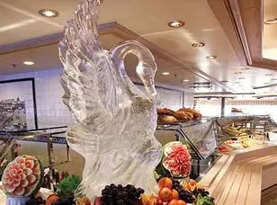 A large ice sculpture of a swan sits in the center of a buffet filled with colorful fruits and vegetables.
