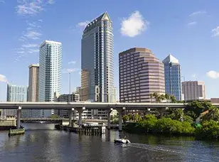 A bustling scene along the Tampa Riverwalk: Pedestrians and cyclists enjoy the waterfront path lined with palm trees, while boats cruise the river and vibrant buildings dot the skyline.