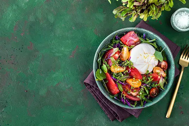 On a green stone background, a salad with tomatoes, arugula, Burrata cheese, and microgreens.