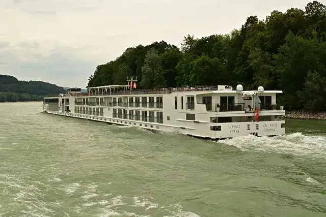 A white river cruise ship named Viking Freya is sailing down a wide river. The ship has multiple decks and is surrounded by lush green hills and trees. The sky is cloudy and the water is choppy.